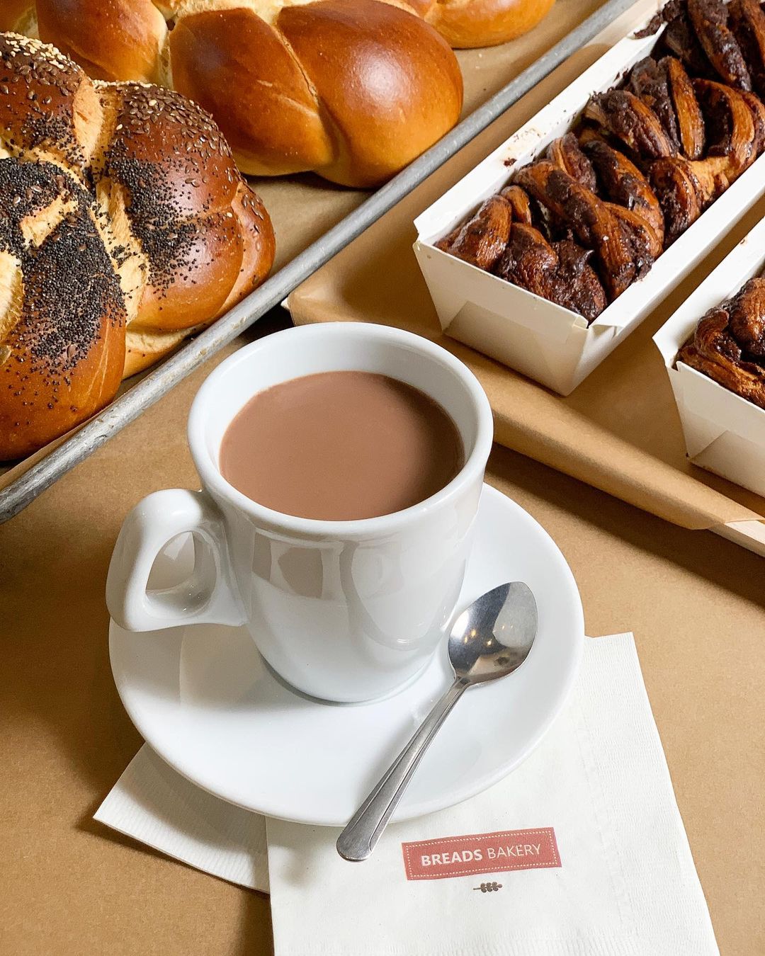 a cup of coffee with a Breads Bakery branded napkin sits in front of a tray of fresh rolls and a tray of chocolate babka