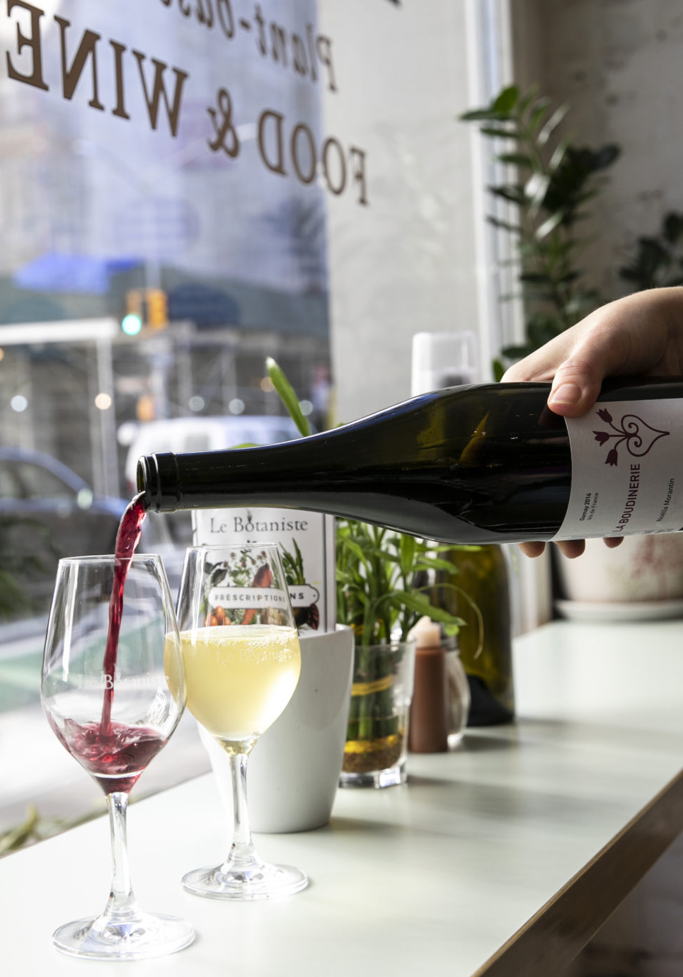 a hand pours a glass of red wine next to a glass of white wine in the window of Le Botaniste