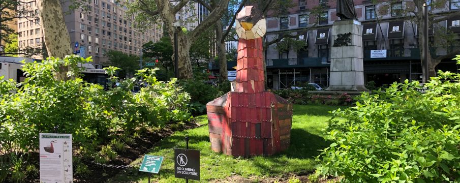 a wooden sculpture of a red necked grebe sits on the grass area of Dante Park