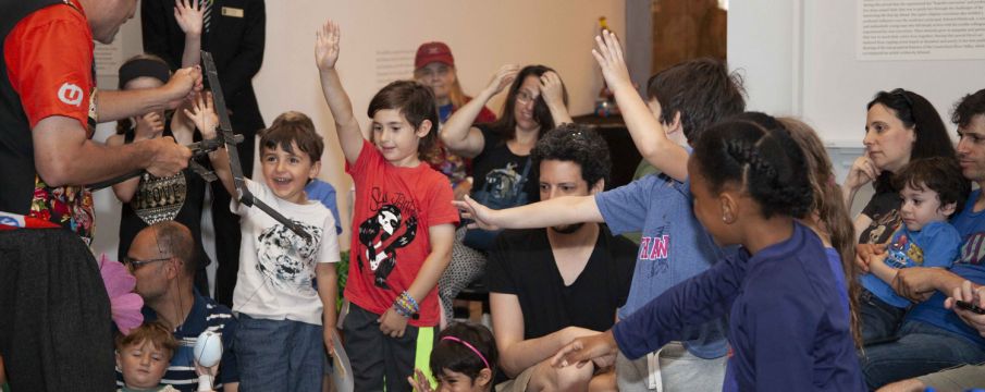 a group of excited children surround an artist and interact with his piece of art that involves wood and string