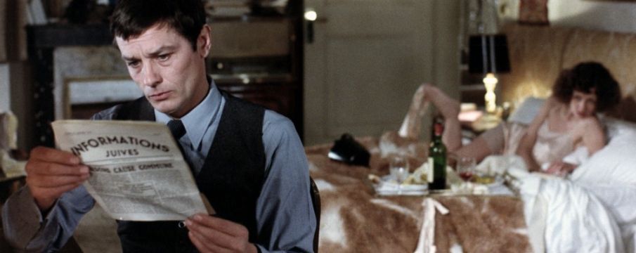 a still from Monsieur Klein--Klein reads the paper while a woman lounges in bed in the background