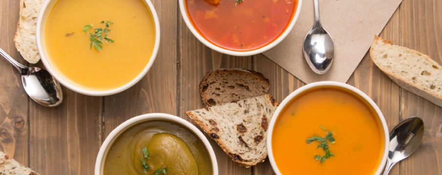 three soups on a wooden cutting board at Le Pain Quotidien with slices of bread and a hand dipping bread into one bowl