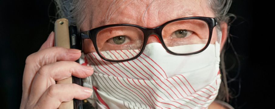 a close up photo of an older woman wearing a face mask with glasses and speaking into her iPhone for a phone call 