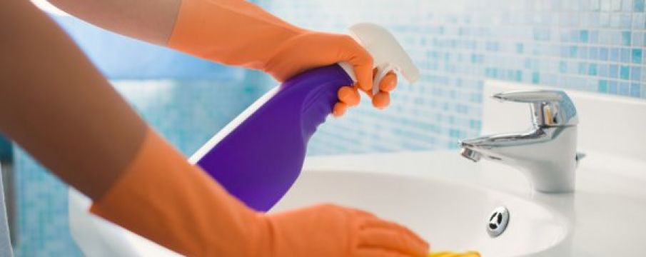 a person's hands wearing rubber cleaning gloves wipes down a sink counter with a rag and cleaning spray