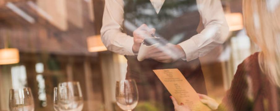 looking through the window, a waiter takes the order of a woman at a table of a nice restaurant with wine glasses on the table