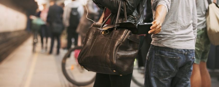 a man's arm pulls a wallet from a woman's bag who is waiting on the subway platform